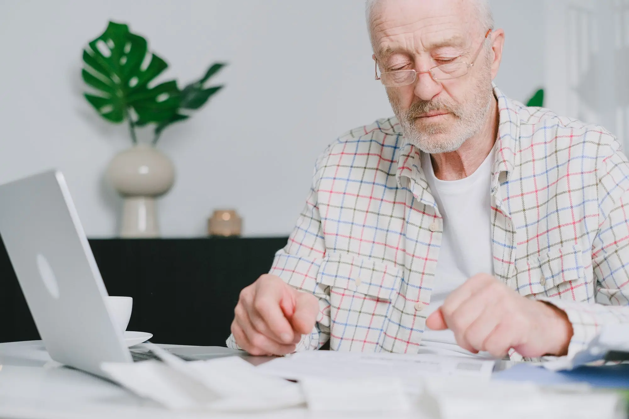 Older business owner in reading glasses going through printed savings and tax statements beside a laptop at home